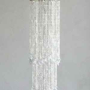 May include: A tiered cylindrical chandelier featuring multiple strands of clear, faceted crystal beads suspended from a circular silver-toned metal frame. The structure hangs from a central point with thin metal wires, creating a cascading effect.
