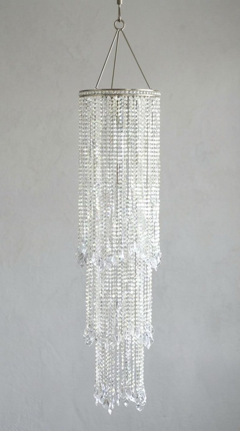 May include: A tiered cylindrical chandelier featuring multiple strands of clear, faceted crystal beads suspended from a circular silver-toned metal frame. The structure hangs from a central point with thin metal wires, creating a cascading effect.