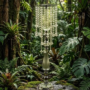 Puede incluir: Un soporte de centro de mesa alto de vidrio transparente con un dise&ntilde;o escalonado y cuentas colgantes facetadas tipo cristal. La estructura incluye una columna central delgada y estriada sobre una base cuadrada para exhibir objetos.