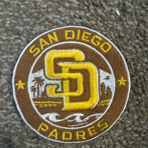 May include: Circular embroidered patch featuring the San Diego Padres logo. The logo includes the letters "SD" in yellow, surrounded by a brown border with the words "San Diego Padres". Palm trees and stars are also included.