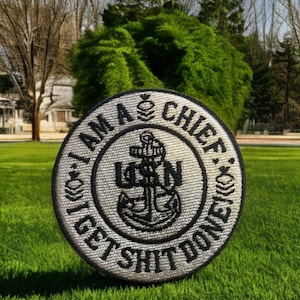 May include: A round, embroidered patch with the text "I AM A CHIEF, I GET SHIT DONE" in black on a silver background. The center features a USN anchor emblem. The patch is set on a green grassy lawn.