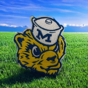 May include: Embroidered patch featuring a yellow bear wearing a white hat with a blue "M" on it. The bear has blue accents and is set against a backdrop of green grass and a blue sky with clouds. The patch is likely for a sports team.