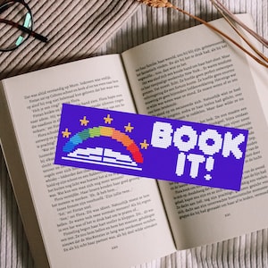 Peut inclure: Un marque-page violet avec un arc-en-ciel, des étoiles et un livre avec le texte "BOOK IT!" en blanc.