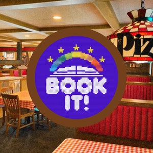 May include: A blue and brown circular sticker with a rainbow, a book, and the text "BOOK IT!" in white. The sticker is on a brown background with a red and white checkered tablecloth in the foreground.