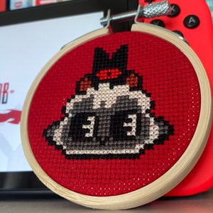 May include: A red and white cross-stitch embroidery of a character from the video game "Cult of the Lamb" in a wooden embroidery hoop.