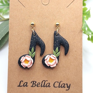 May include: A pair of black glitter musical note earrings with pink flowers and green leaves. The earrings are on a brown card with the text "La Bella Clay".