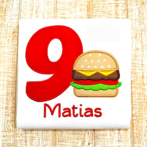 May include: Red number nine with a cartoon burger and the name Matias on a white t-shirt.