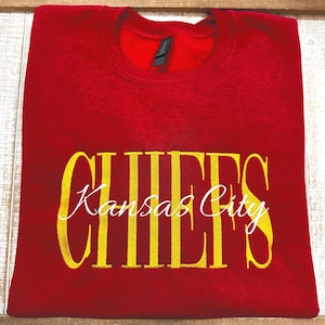 May include: A red crewneck sweatshirt featuring the word CHIEFS embroidered in large yellow block letters across the chest, with the words Kansas City written in a white cursive script font layered over the yellow text on the front.
