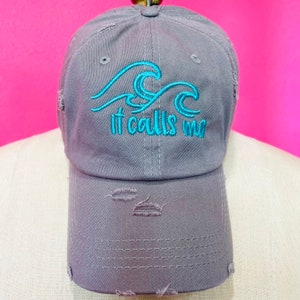May include: Gray baseball cap with distressed details. The cap features embroidered turquoise waves and the phrase "it calls me". A stylish accessory for outdoor activities.