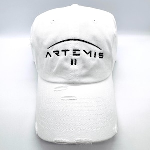 May include: White baseball cap with distressed details. The cap features the embroidered text "ARTEMIS II" in black, arched across the front. The hat has a curved brim and an adjustable strap at the back.