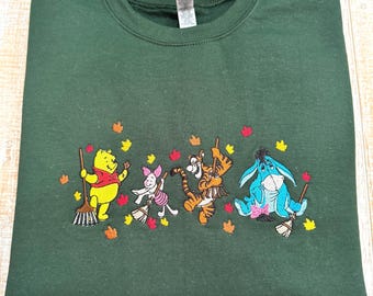 May include: A dark green crewneck sweatshirt featuring an embroidered design of Winnie the Pooh, Piglet, Tigger, and Eeyore. Each character is holding a broom, surrounded by small, colorful embroidered autumn leaves in shades of red, orange, and yellow.