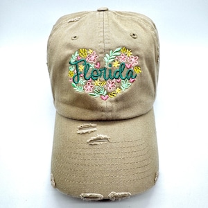 May include: A tan distressed baseball cap with a green embroidered heart featuring the word "Florida" and colorful flowers.