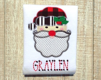 May include: White long-sleeve shirt with an applique of a Santa Claus face. The Santa has a red and black plaid hat, a black and white striped hat band, a white beard with black polka dots, and a red nose. The name "GRAYLEN" is embroidered in red below the Santa.