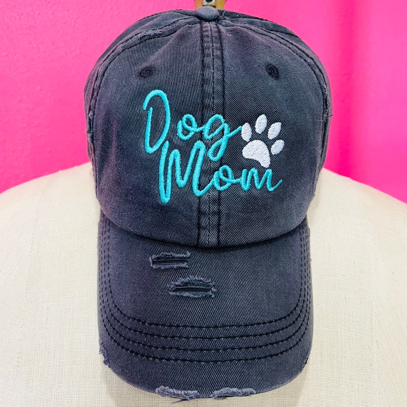 May include: Distressed navy blue baseball cap with the embroidered text "Dog Mom" in light blue script and a white paw print. The cap has a curved brim and distressed details for a vintage look. A stylish accessory for dog lovers.