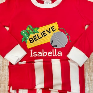 May include: Red and white striped pajamas with long sleeves and a red top. The top has an embroidered design of a yellow sign that says "Believe" with a green bow and a silver bell. The name "Isabella" is embroidered below the sign.