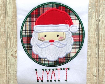 May include: White Christmas-themed baby onesie with a red and green plaid circle featuring a white embroidered Santa face and the name "WYATT" embroidered below.