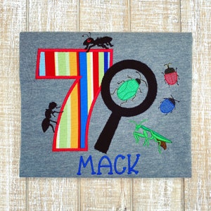 May include: A gray t-shirt with a colorful striped number 7, a magnifying glass, and various bugs embroidered on it. The text "MACK" is embroidered below the design.