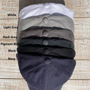 May include: A stack of six baseball caps in various colors: white, light grey, dark grey, pigment black, black, and navy. Each cap has a rounded crown and a button on top. The colors are labeled in white text.