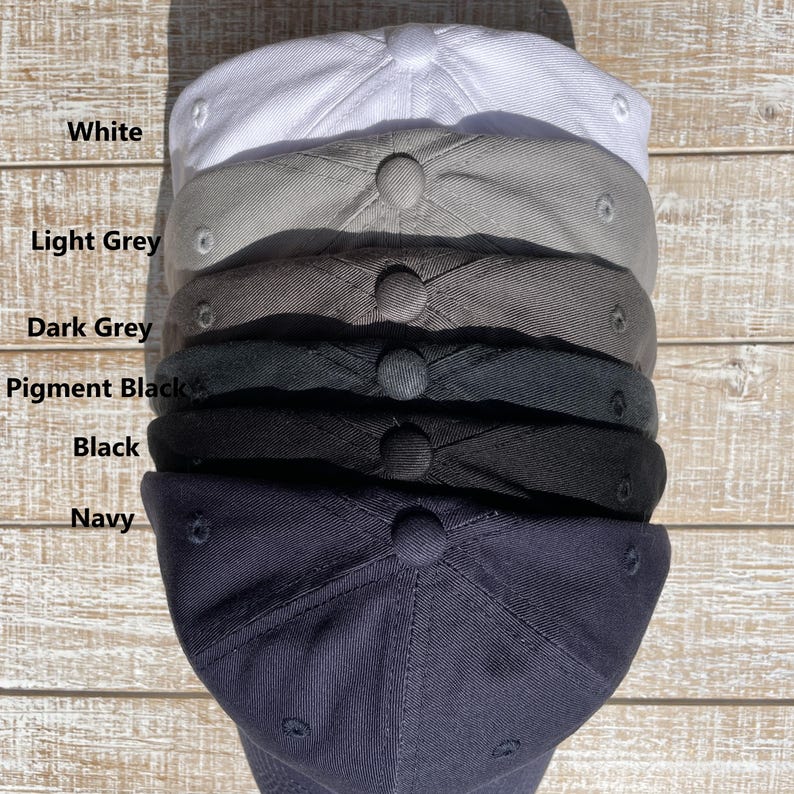 May include: A stack of six baseball caps in various colors: white, light grey, dark grey, pigment black, black, and navy. Each cap has a rounded crown and a button on top. The colors are labeled in white text.