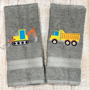 May include: Two gray hand towels with embroidered images of construction vehicles. One towel features a yellow excavator with a black track and the other towel features a yellow dump truck with black wheels.