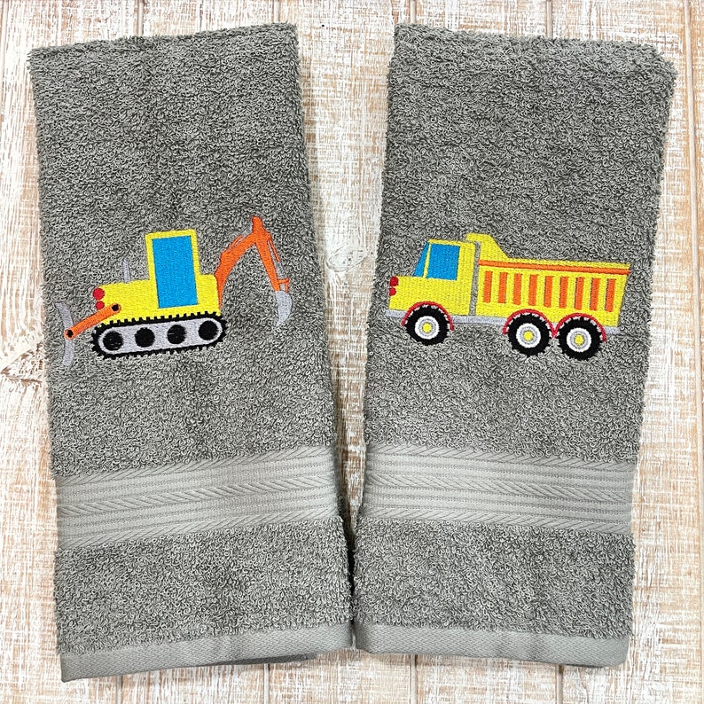 May include: Two gray hand towels with embroidered images of construction vehicles. One towel features a yellow excavator with a black track and the other towel features a yellow dump truck with black wheels.