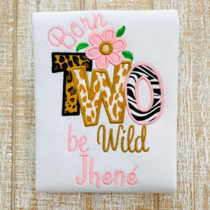 Peut inclure: Une grenouill&egrave;re blanche avec les mots "Born Two Be Wild" brod&eacute;s en rose et or. Le chiffre deux est d&eacute;cor&eacute; d'un imprim&eacute; l&eacute;opard et z&egrave;bre. La grenouill&egrave;re est personnalis&eacute;e avec le nom "Jhen&eacute;" en rose.