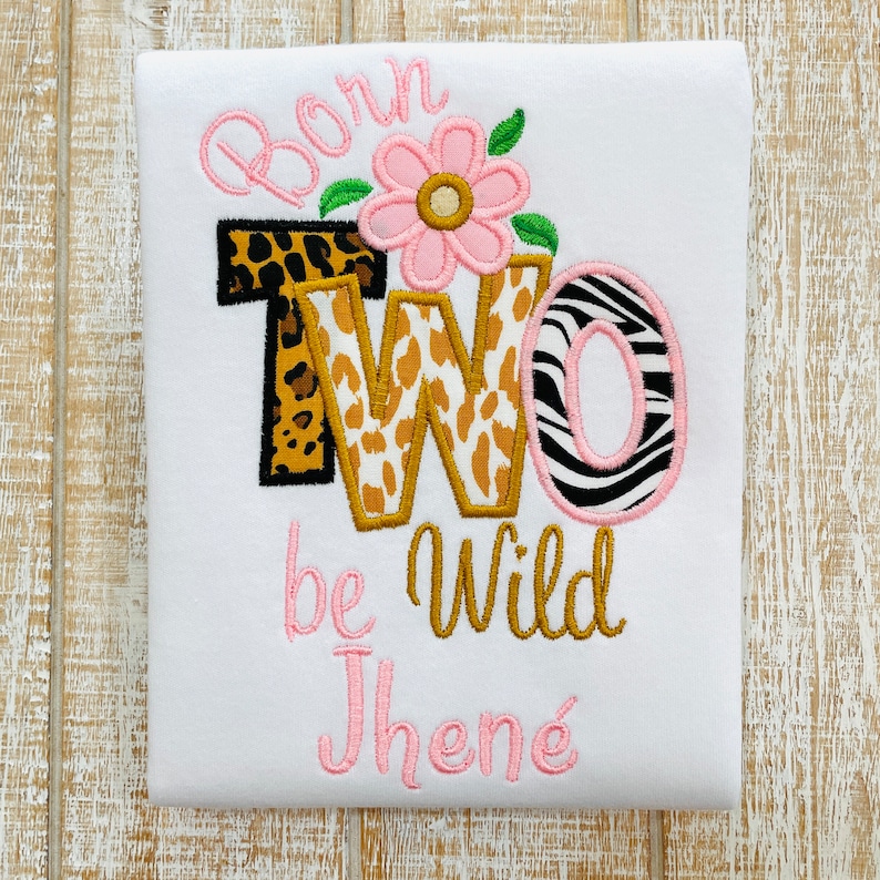 Peut inclure: Une grenouill&egrave;re blanche avec les mots "Born Two Be Wild" brod&eacute;s en rose et or. Le chiffre deux est d&eacute;cor&eacute; d'un imprim&eacute; l&eacute;opard et z&egrave;bre. La grenouill&egrave;re est personnalis&eacute;e avec le nom "Jhen&eacute;" en rose.