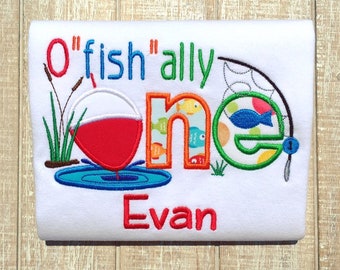May include: A white onesie with the text "O'fish'ally one" in colorful embroidered letters. The onesie also features a red and white fishing bobber, a fishing rod, and the name "Evan" embroidered in red.