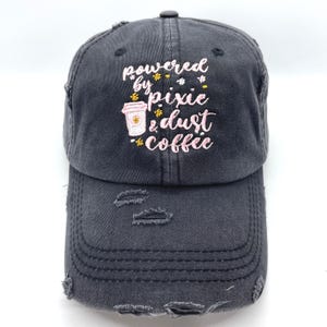 May include: Black distressed baseball cap with embroidered text "powered by pixie dust coffee" in pink and yellow. The cap features a small embroidered image of a coffee cup.