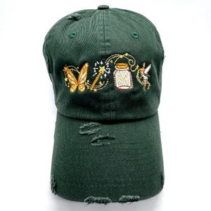 May include: A dark forest green baseball cap with a distressed, worn-in finish. The front features detailed embroidery of a golden butterfly, a magic wand, a glowing jar, and a small fairy figure, all rendered in shades of gold, white, and brown.
