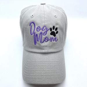 May include: A gray baseball cap with the embroidered text "Dog Mom" in purple script and a black paw print. The cap has a curved brim and an adjustable strap.