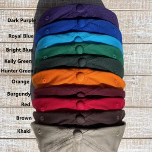 May include: A stack of baseball caps in various colors, including dark purple, royal blue, bright blue, kelly green, hunter green, orange, burgundy, red, brown, and khaki. Each cap has a button on top.