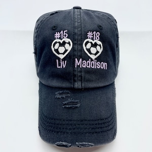 May include: Black distressed baseball cap with two soccer ball heart designs, the numbers #15 and #18, and the names Liv and Maddison embroidered in purple.