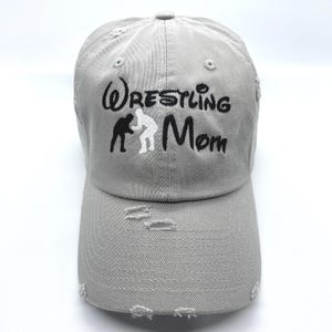 May include: A gray, distressed baseball cap with the words "Wrestling Mom" embroidered in black. The design includes a silhouette of two wrestlers. The cap has a curved brim and adjustable strap.