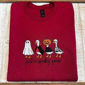 Puede incluir: Una camiseta granate con un dise&ntilde;o bordado de cuatro gansos disfrazados para Halloween: un fantasma, un vampiro, una calabaza y un esqueleto. El texto "just a spooky goose" est&aacute; debajo.