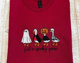 May include: A dark red crew neck t-shirt featuring an embroidered design of four geese dressed in Halloween costumes: a ghost, a vampire, a pumpkin-headed goose, and a skeleton. Below the geese, the text reads, just a spooky goose.