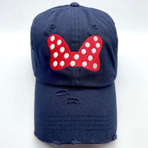 May include: A navy blue baseball cap with a red and white polka dot bow embroidered on the front. The cap has distressed detailing.