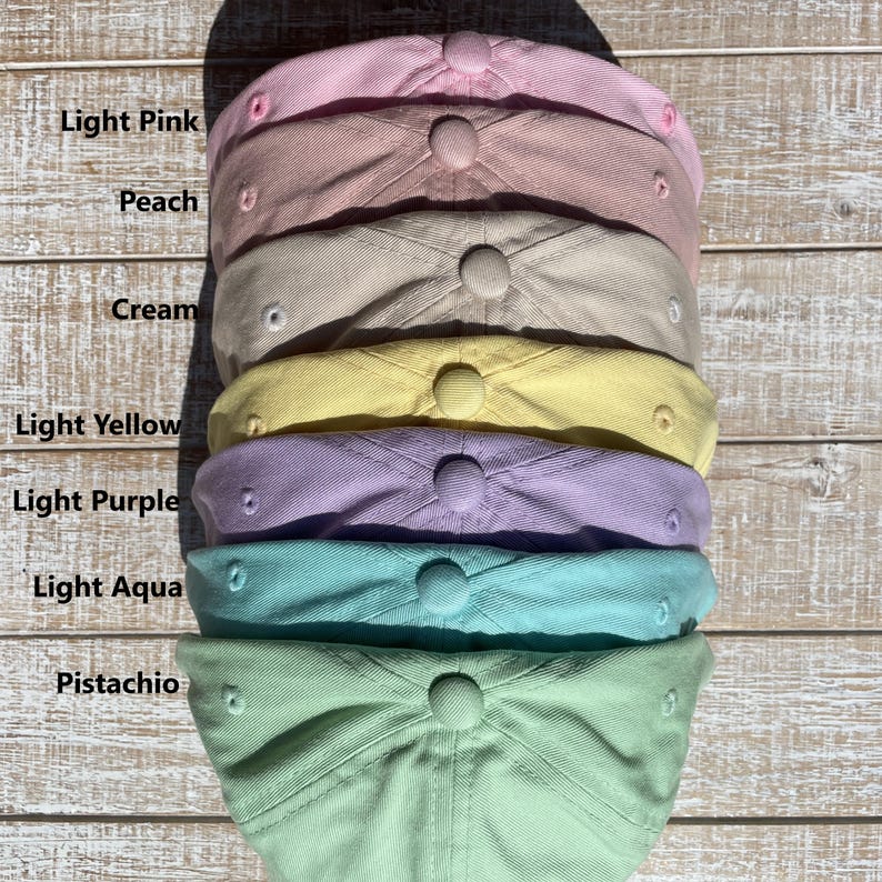 May include: A stack of pastel-colored baseball caps, including light pink, peach, cream, light yellow, light purple, light aqua, and pistachio. Each cap has a curved brim and a button on top. The colors are labeled on the left side of the image.