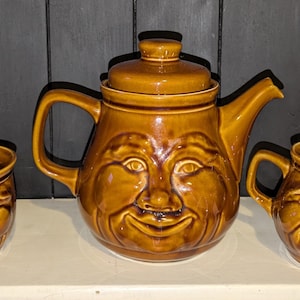 May include: A brown ceramic teapot, sugar bowl, and creamer set with whimsical faces. Each piece features a smiling face with brown eyes and a wide mouth.