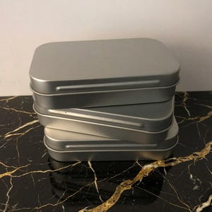 May include: A stack of three rectangular silver metal tins with rounded corners and fitted lids. Each container features a smooth, matte finish and a simple, functional design. The text Backyardsounds is visible in the bottom right corner.