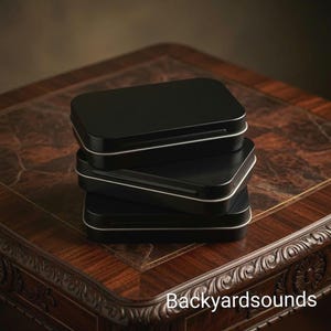 May include: A stack of three rectangular black metal tins with rounded corners and silver-colored rims. The tins are stacked vertically, each featuring a smooth, matte black finish on the lid and base, with the text Backyardsounds visible.