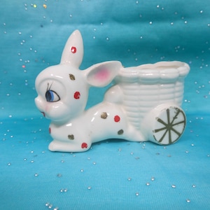 May include: A white ceramic bunny planter with a basket and wheel design. The bunny has blue eyes, pink ears, and red and brown spots. The planter is on a blue background.