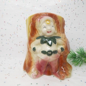May include: A vintage ceramic planter shaped like a tree stump, featuring a smiling elf figure. The elf wears a white outfit with a green collar and belt, and pink pants. The planter is painted in shades of brown, yellow, and pink.