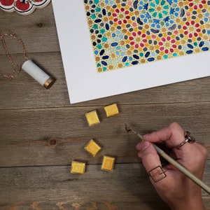 Op de afbeelding: Een bovenaanzicht van een houten oppervlak met een kleurrijke geometrische kunstdruk, gouden aquarelverf en een ketting. Een hand houdt een kalligrafiepen vast. De kunstdruk heeft een gouden rand.