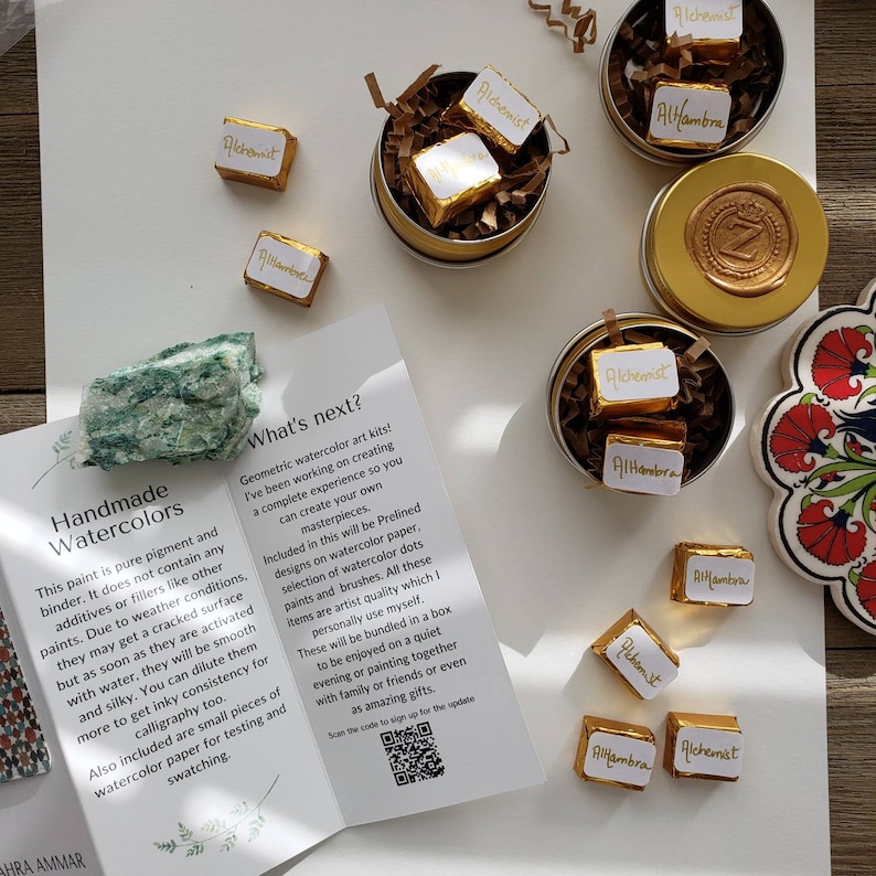 Op de afbeelding: Bovenaanzicht van handgemaakte aquarel kunstkits. De afbeelding toont kleine gouden blikken met chocolaatjes met het opschrift "Alchemist" en "Alhambra", een groene steen en een brochure die de kits beschrijft. De kits bevatten voorgelijnde ontwerpen, aquarelpunten en penselen.