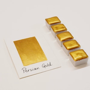Op de afbeelding: Een verzameling van vijf vierkante, goudkleurige aquarelverfblokjes met witte bases, gerangschikt naast een witte kaart met een gouden verfstaal en de handgeschreven tekst "Persian Gold". De achtergrond is wit.