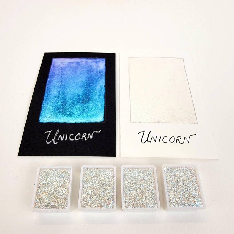 May include: A collection of watercolor paints and swatches. The swatches are on black and white paper, with the word "Unicorn" written in cursive. Four small, rectangular containers hold glittery paint.