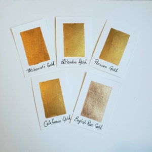 Op de afbeelding: Vijf gouden aquarelverfstalen op witte kaarten, met de namen "Alchemist's Gold", "Alhambra Gold", "Persian Gold", "California Gold" en "English Rose Gold". De gouden tinten vari&euml;ren van helder tot zacht roosgoud.
