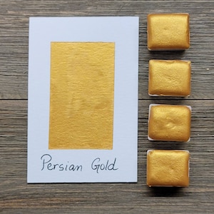 Op de afbeelding: Een witte kaart met een rechthoekige goudverfstaal en de tekst "Persian Gold". Vier vierkante goudverfpotjes staan rechts van de kaart. De achtergrond is een houten oppervlak.