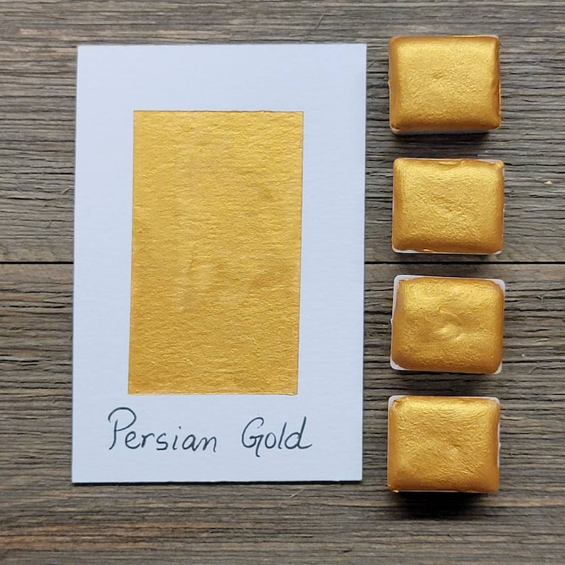 Op de afbeelding: Een witte kaart met een rechthoekige goudverfstaal en de tekst "Persian Gold". Vier vierkante goudverfpotjes staan rechts van de kaart. De achtergrond is een houten oppervlak.